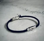 نمای کامل دستبند لنگر نقره با بند نخی مشکی و قفل مگنتی روی سطح سنگی