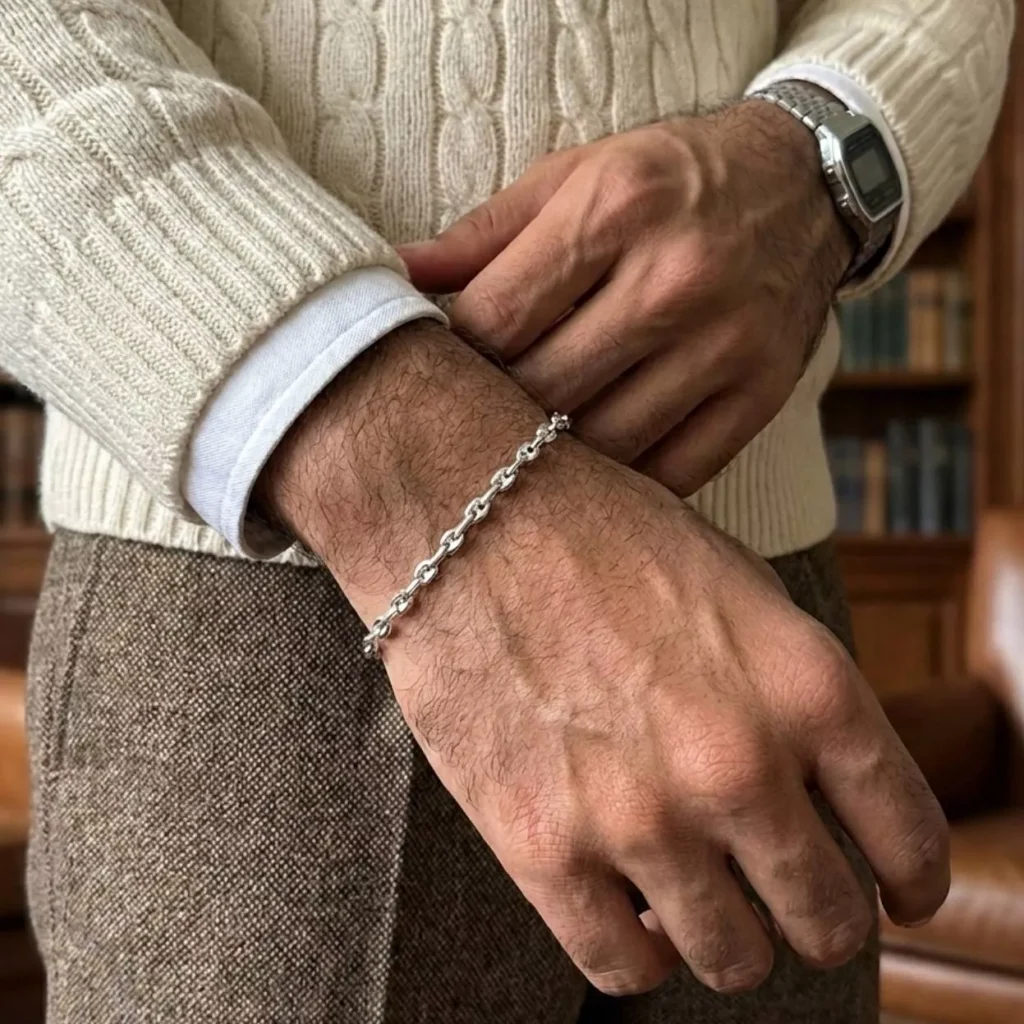 مردی با استایل کلاسیک و پلیور بافت که دستبند نقره زنجیری طرح مارینر را در کنار ساعت مچی بسته است، یک هدیه لوکس برای آقایان.